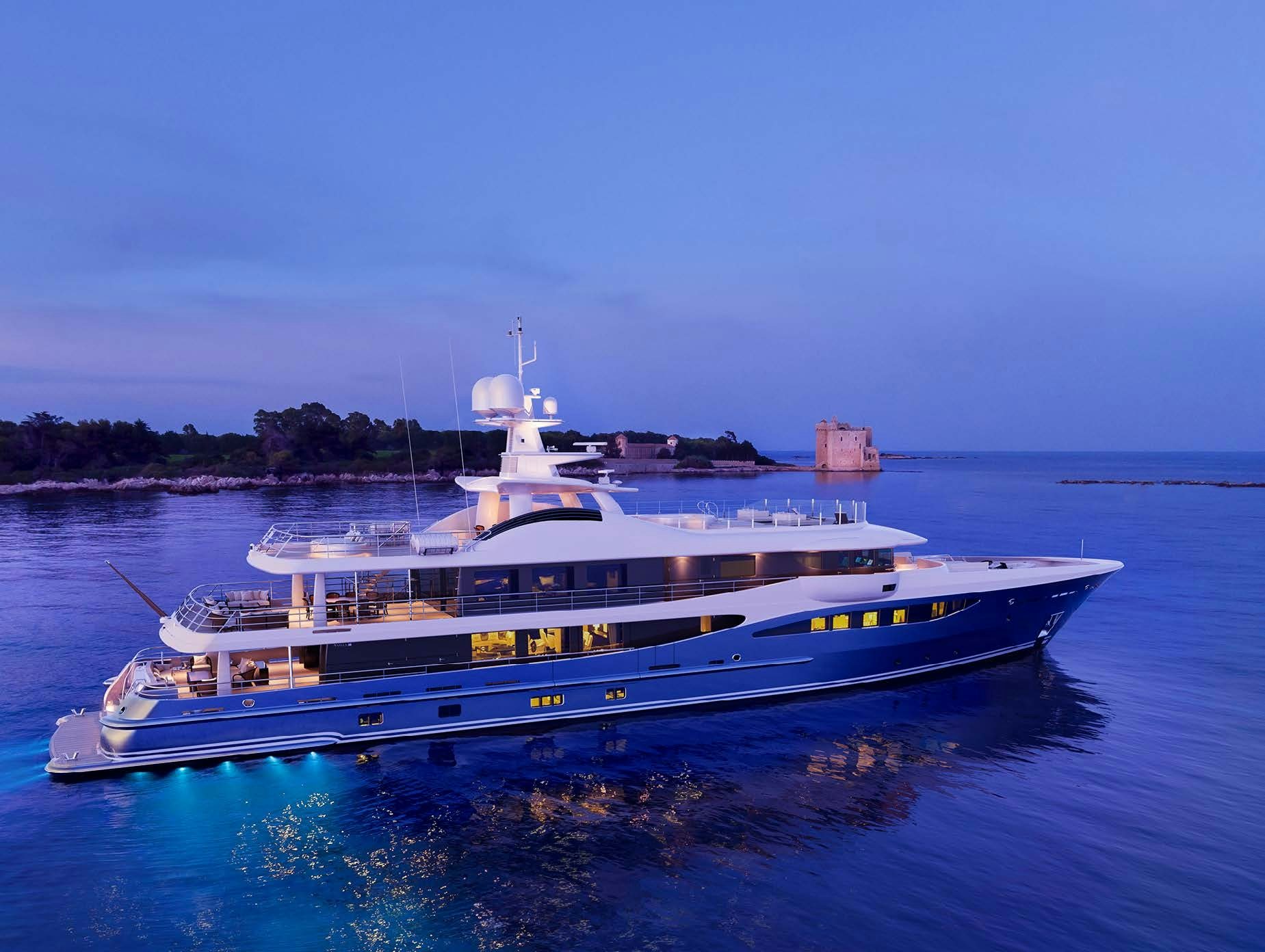 An Amels 180 Superyacht anchored near the coastline at dusk, its lights reflecting on calm waters.