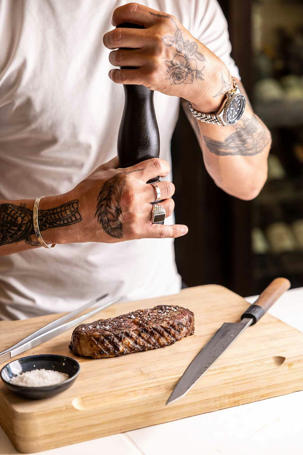 A grilled steak resting on a wooden chopping board being salted by a chef with tattooed arms