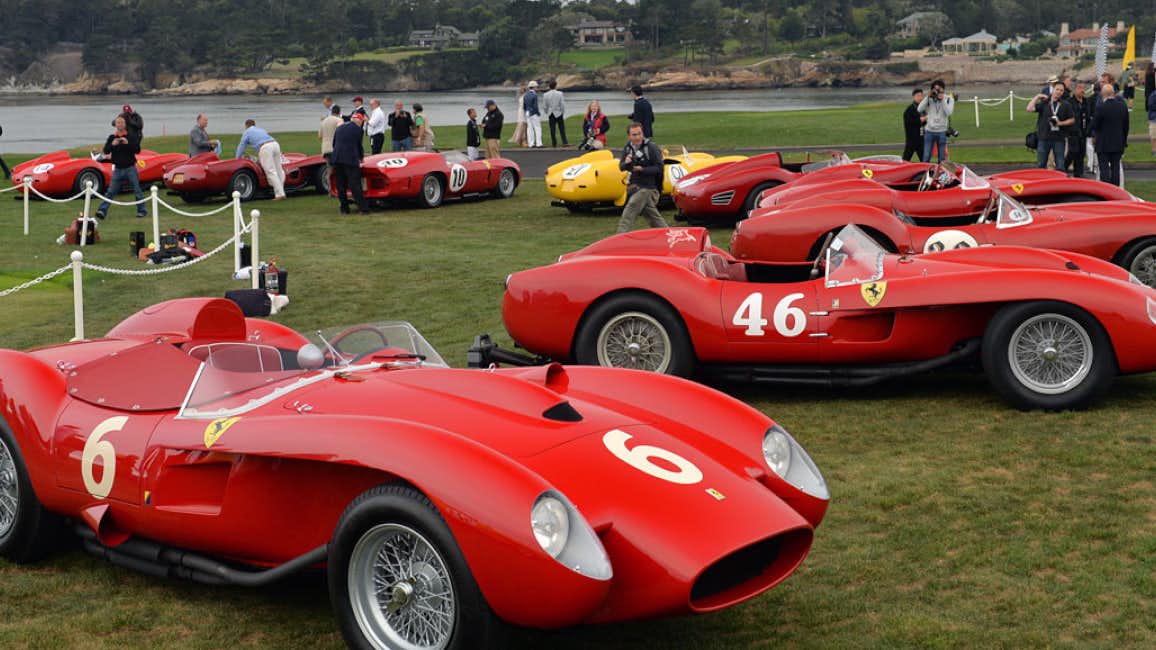A mesmerizing scene featuring a collection of stunning Ferraris and classic cars on a coastal grassland. Enthusiastic car lovers admire these automotive treasures on a cloudy day during the Pebble Beach Concours d'Elegance, a highlight of the Pebble Beach Automotive Week.
