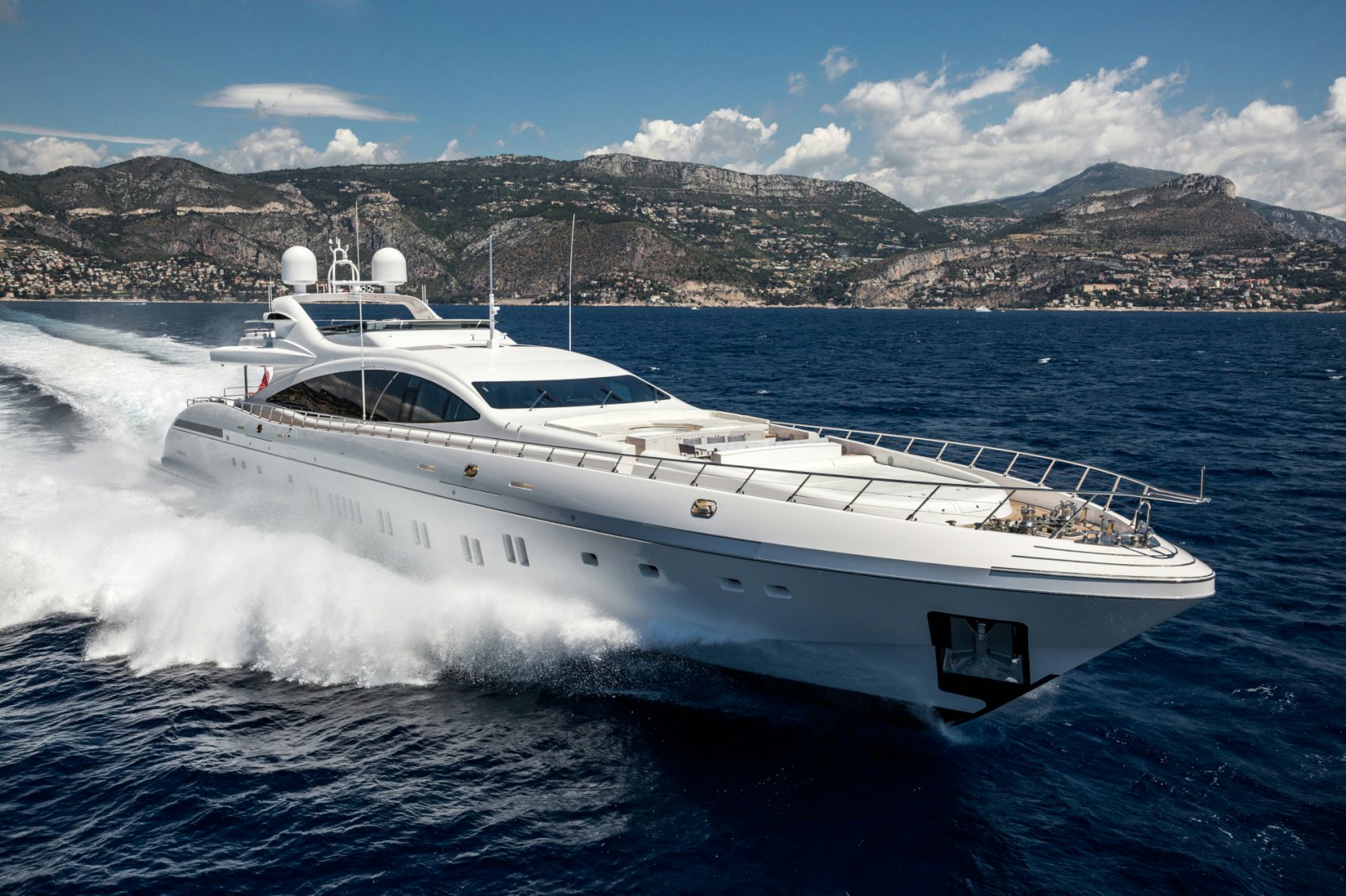 Dynamic Elegance: The Mangusta 165 in Full Sail The Mangusta 165 yacht cutting through the blue waters, with a mountainous coastline in the background under a clear sky.