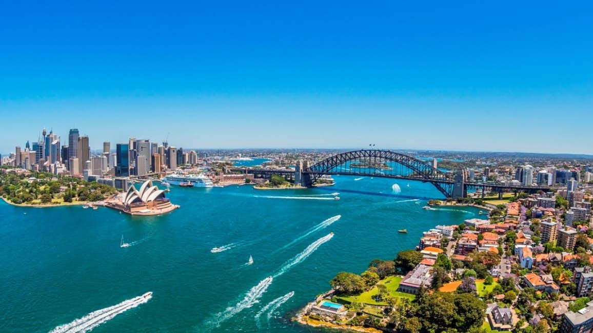 A breathtaking aerial view of Sydney Harbor Bridge against a backdrop of azure skies and the tranquil blue sea. Boats cruise beneath the iconic bridge during the Sydney International Boat Show.