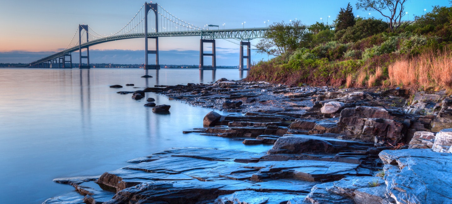 A captivating photo of the Newport Bridge and a rocky beach against a cloudy sunset backdrop during the Newport Charter Show.