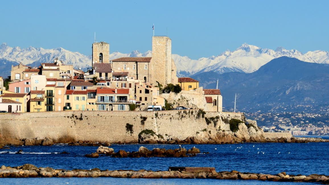 A picturesque side view of the historic Antibes Old Village against the backdrop of the sparkling sea on a sunny and wavy day.