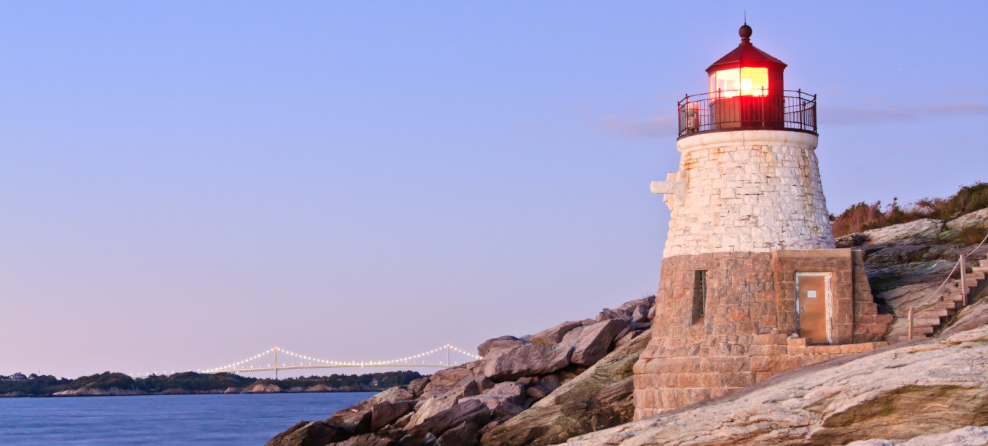 A picturesque view of the Newport Lighthouse perched on a cliff, bathed in the warm glow of the sunset, with the azure sea as the backdrop during the Newport Brokerage Show.