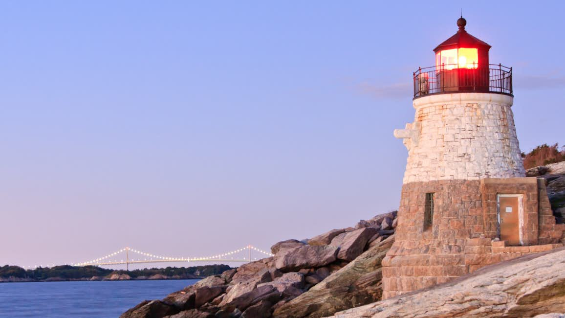 A picturesque view of the Newport Lighthouse perched on a cliff, bathed in the warm glow of the sunset, with the azure sea as the backdrop during the Newport Brokerage Show.