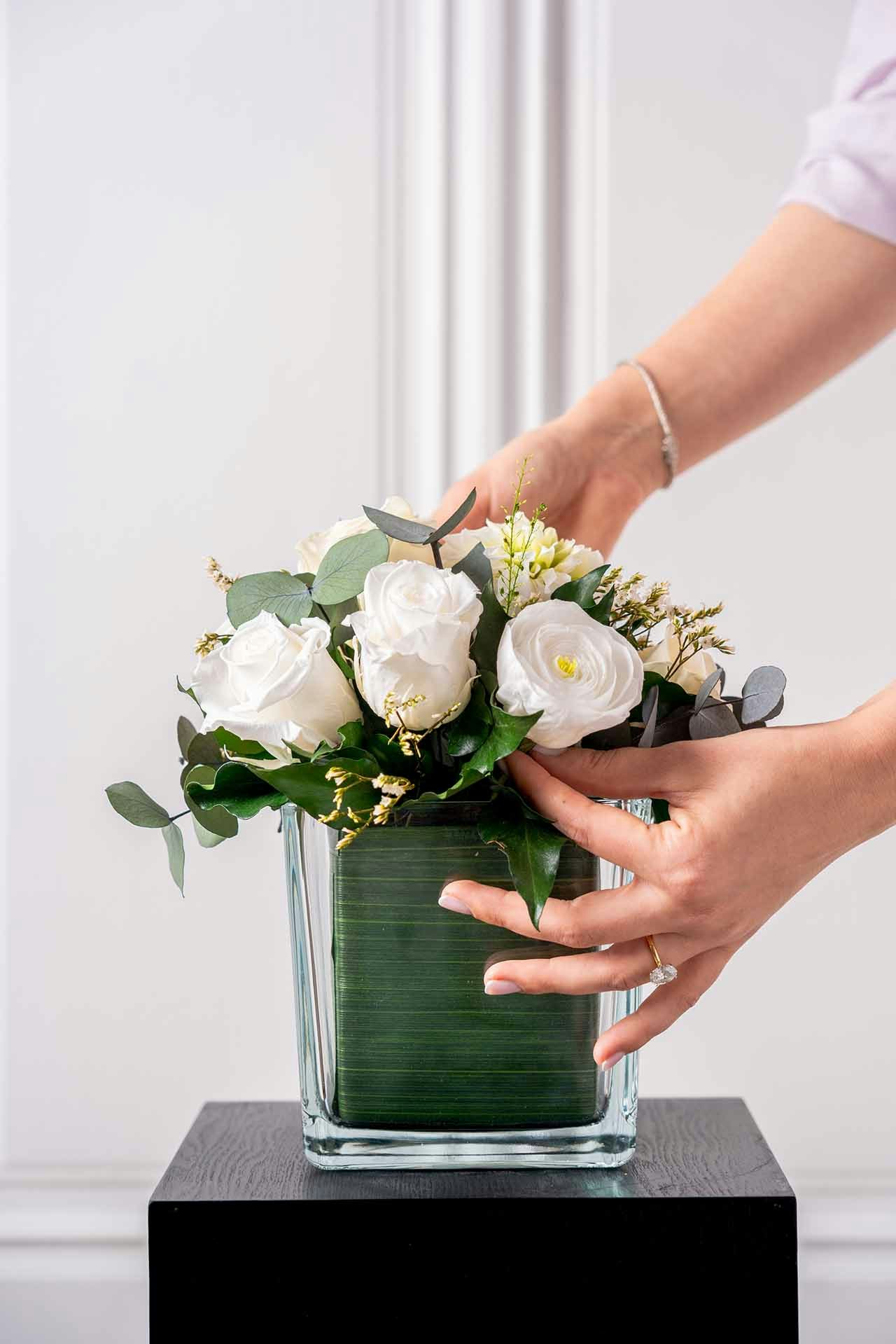 A white bouquet of flowers in a square glass vase with hands adjusting the stems
