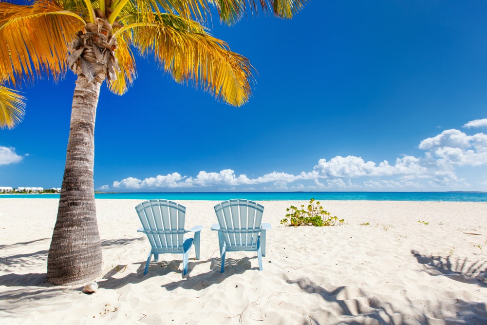 An idyllic scene in Anguilla, featuring a sun-kissed beach with super white sand, two sunbeds under a palm tree—an ideal spot to relax and soak up the sun during your yacht charter in this amazing destination.