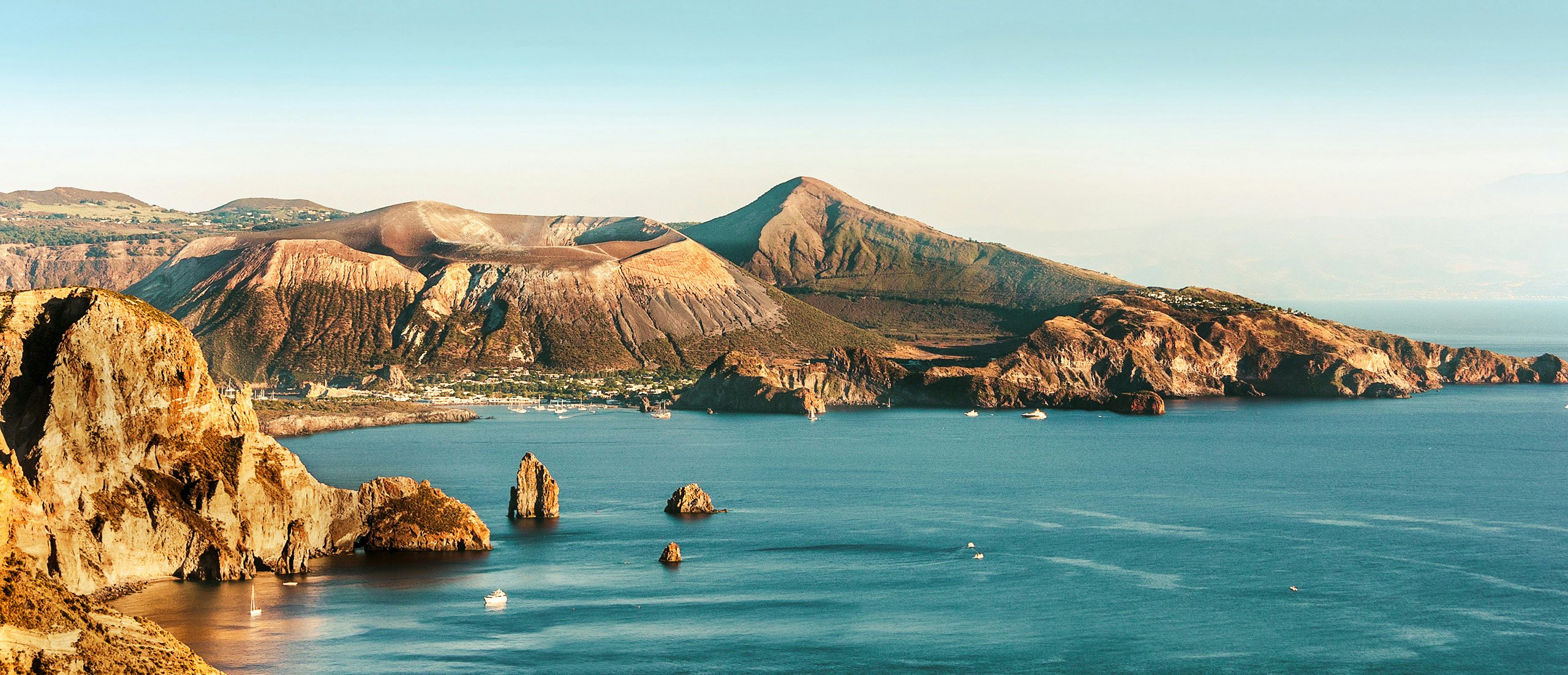 Sun-Kissed Lipari Serenity - Aeolian Islands Yachting with Northrop & Johnson Sunlit Aeolian coastline in Lipari with barren hills and a clear blue sea speckled with boats.