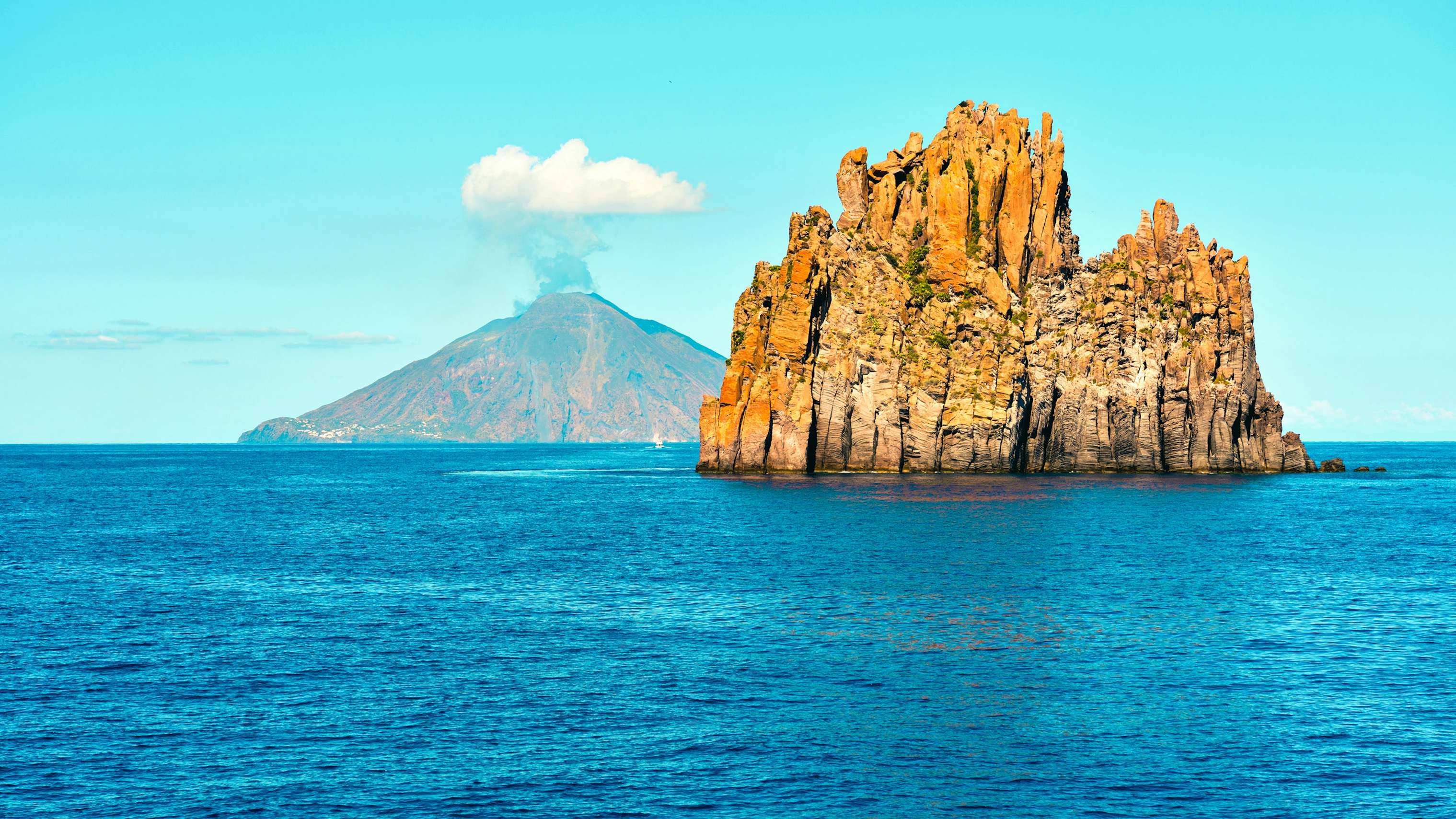 Majestic rock formations rise from the sea with the smoking Stromboli volcano in the background.