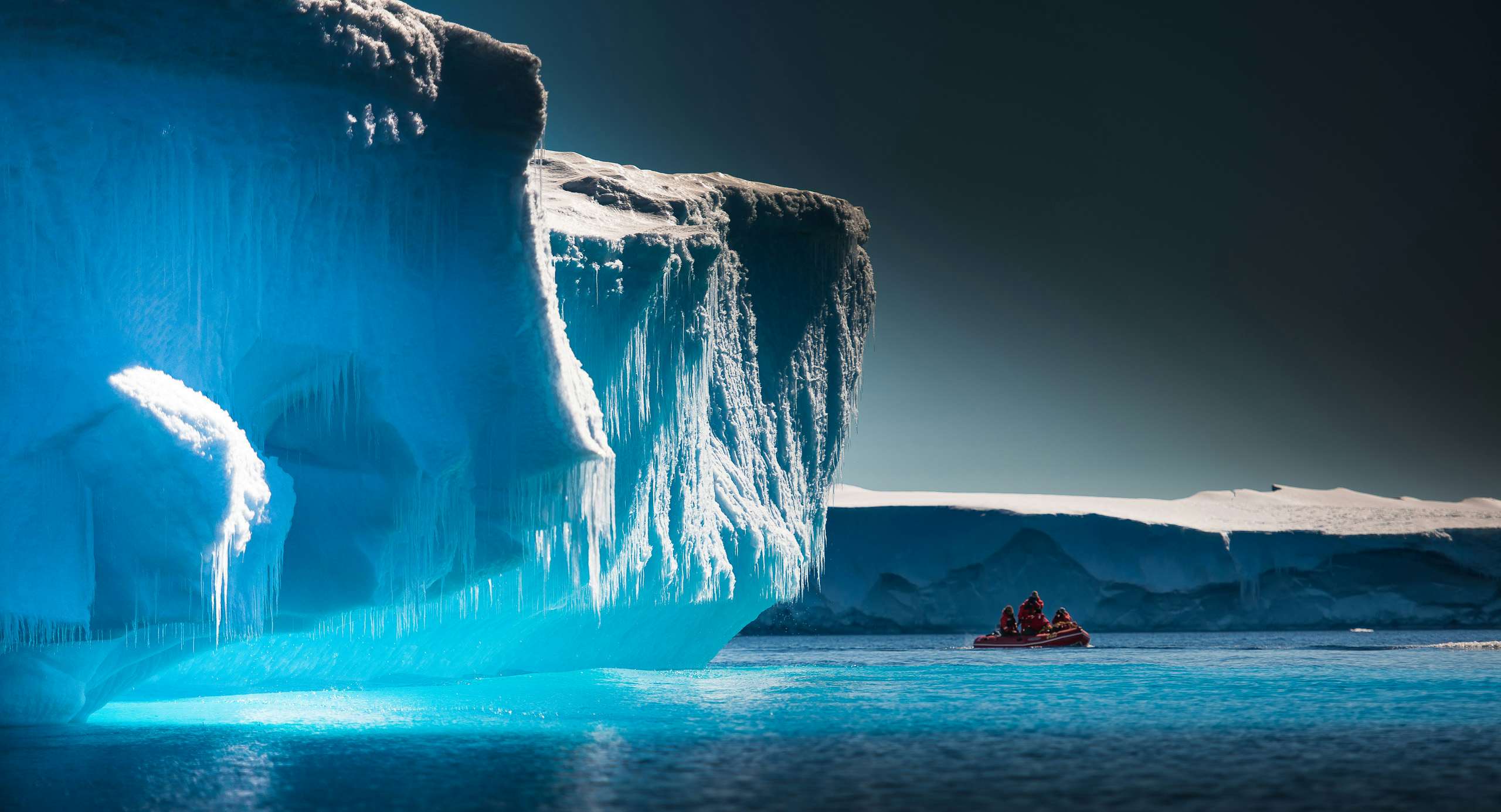 A small boat navigating the icy waters near a massive blue iceberg under a polar sky.