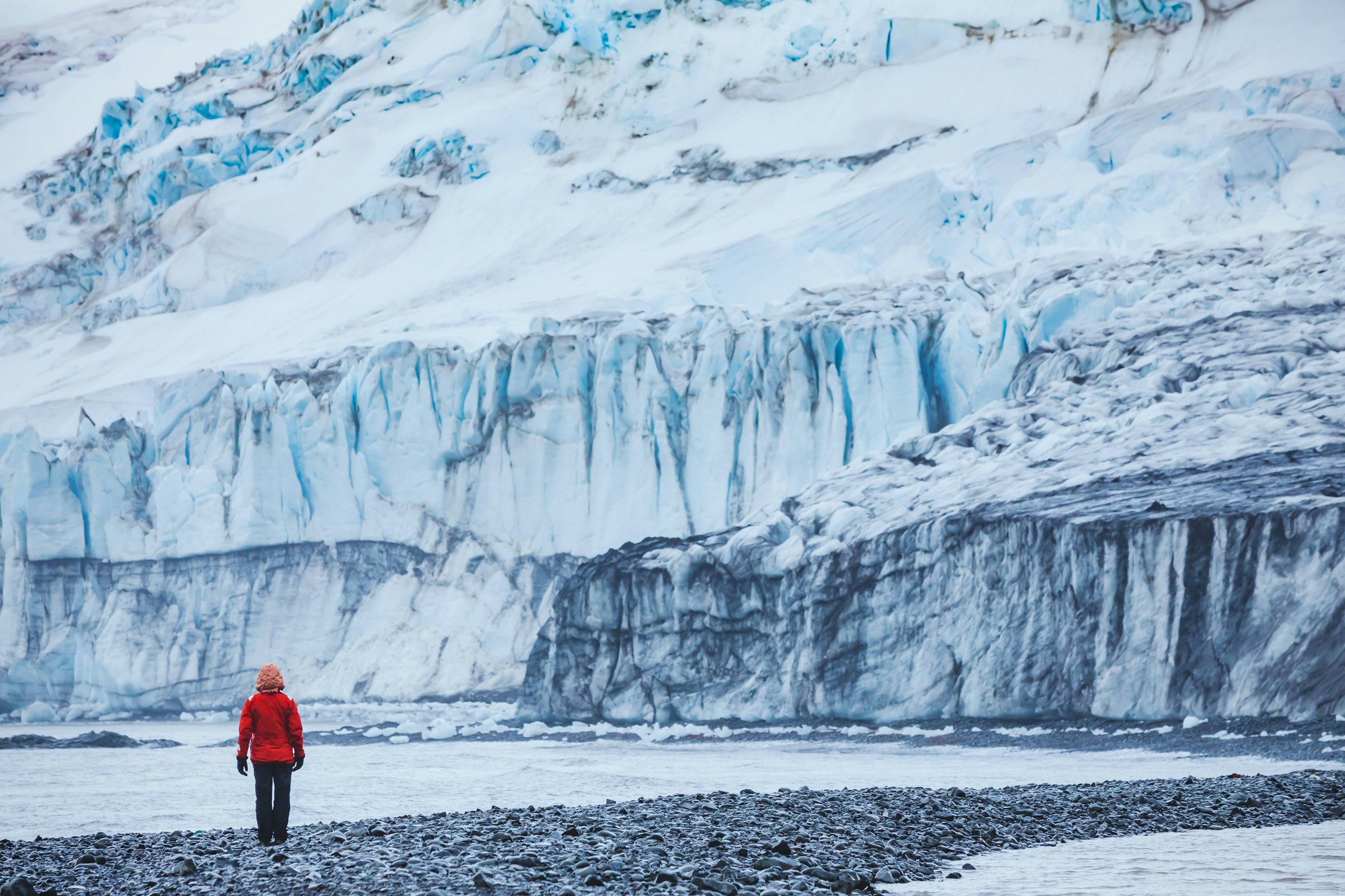 A lone explorer in a red jacket stands before a vast Antarctic glacier.