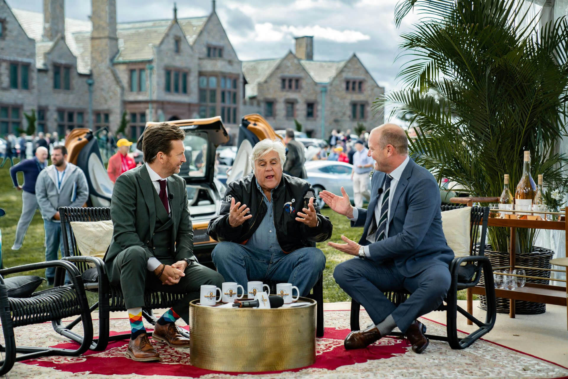 Jay Leno sitting and talking with two men at the Gathering at Audrain&#039;s Newport Concours Motor Week