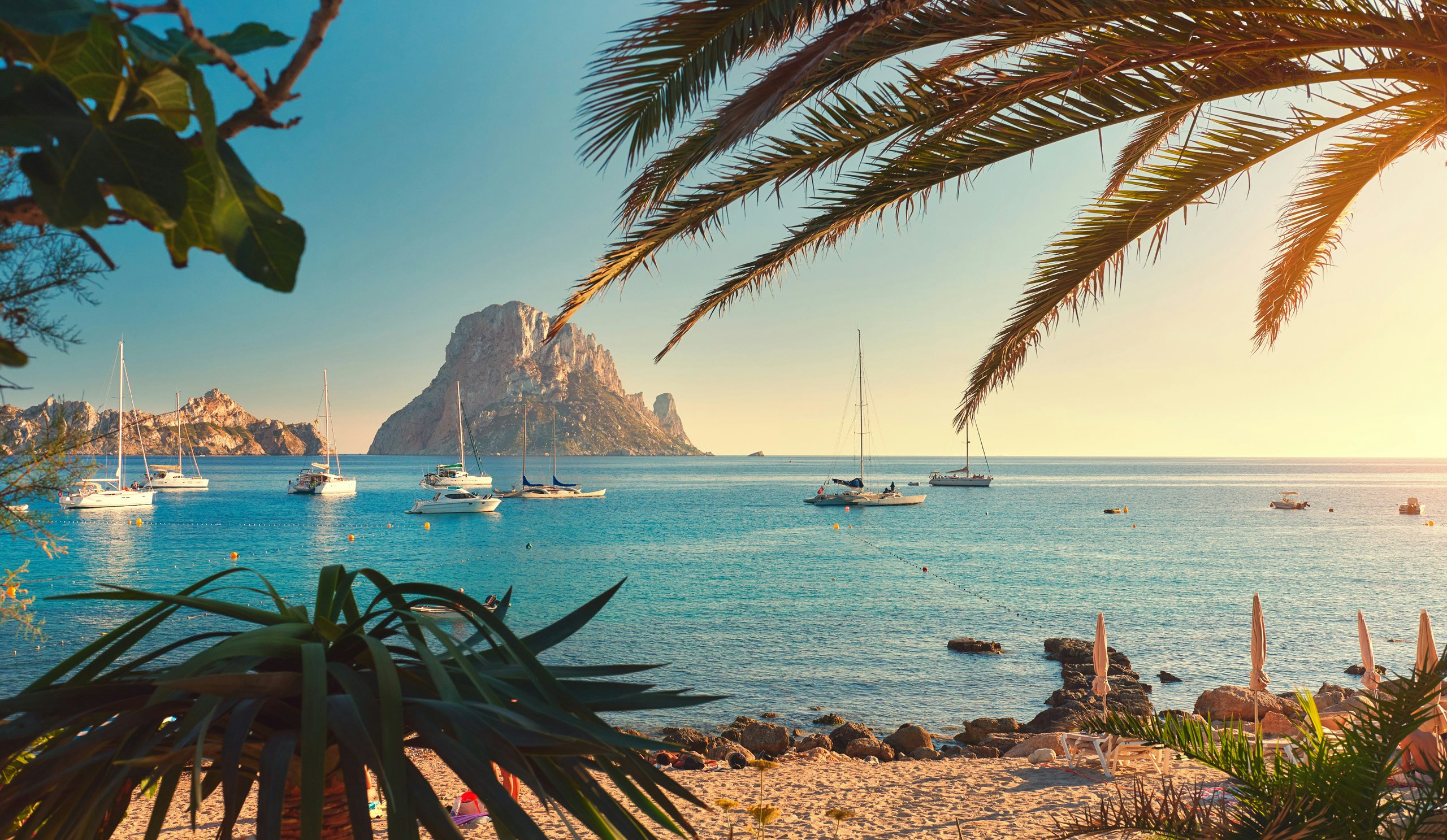 Ibiza's Mystical Horizons | Balearic Yacht Charter | Northrop & Johnson Palm fronds frame a serene view of anchored yachts near the mystical rock of Es Vedrà, off the coast of Ibiza.