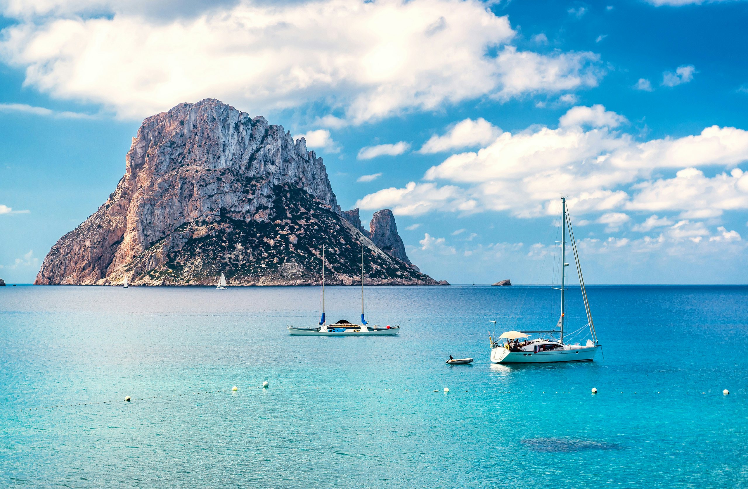 Sailboats glide near the majestic Isla Vedra, rising sharply from the Mediterranean's azure waters.