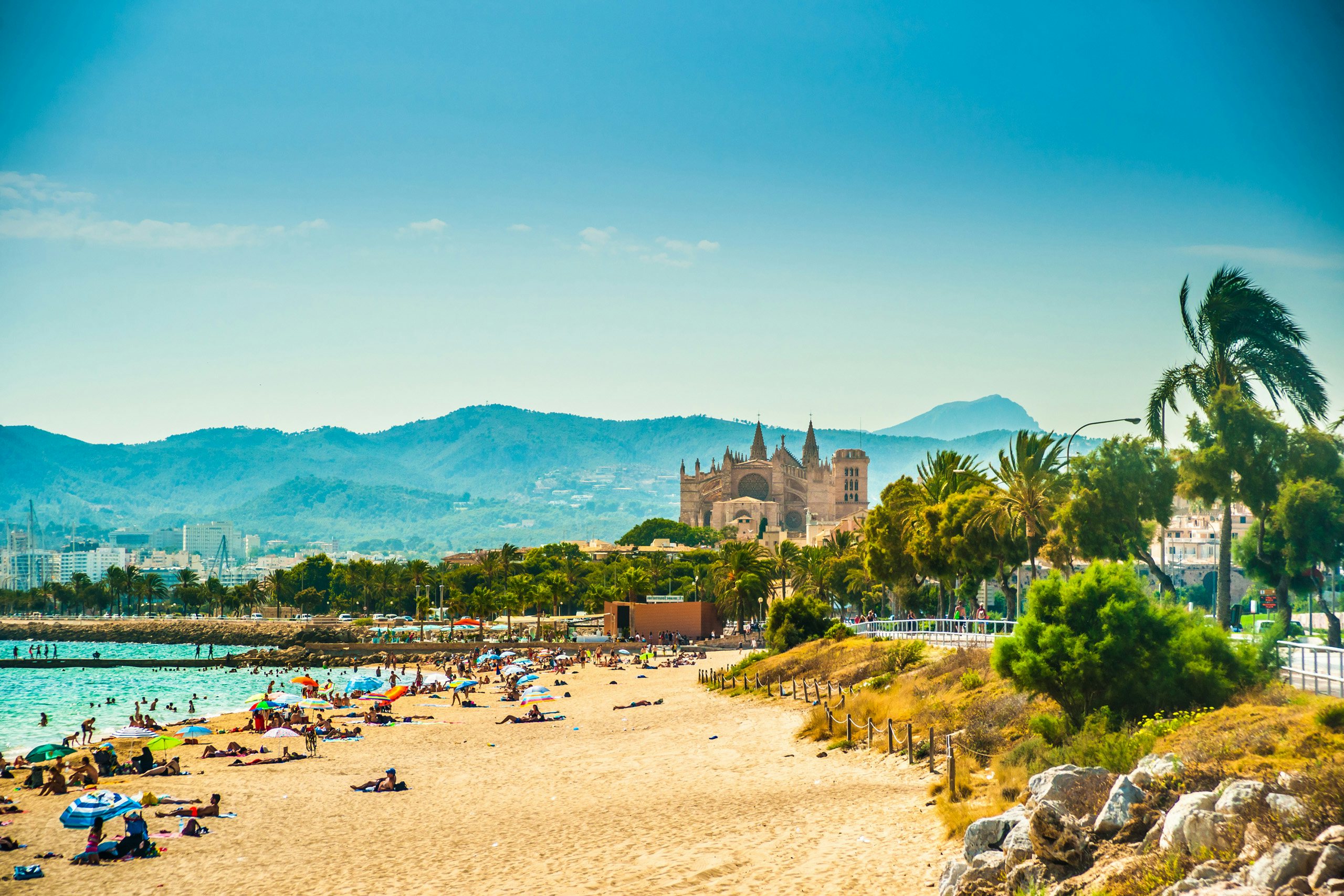 Sandy beach and crystal-clear waters with Palma de Mallorca's historic cathedral in the background.