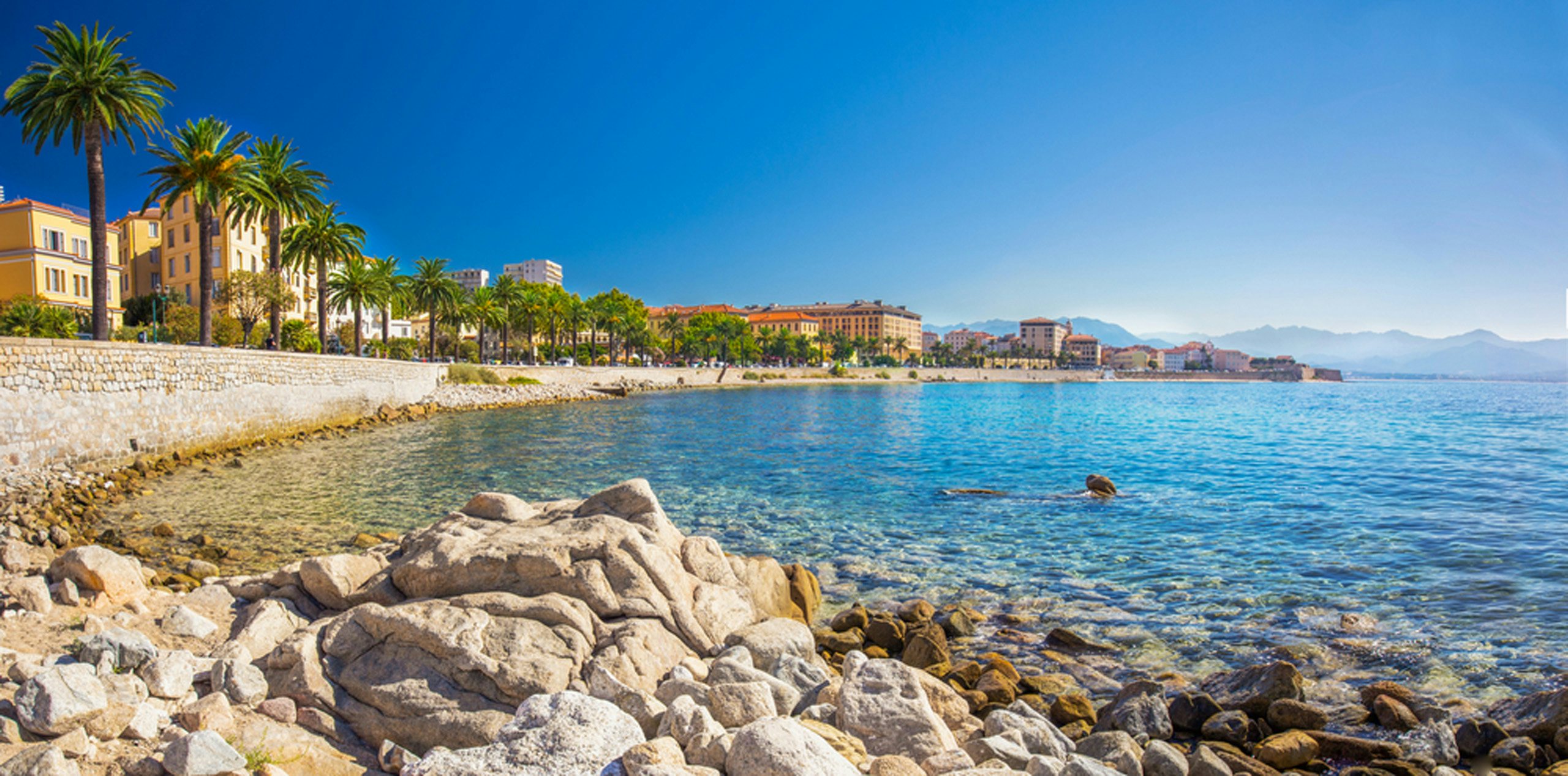 Ajaccio's Rocky Shores | Yacht Charter Escapes | Northrop & Johnson Clear blue waters wash over the rocky beaches of Ajaccio, Corsica, with vibrant city architecture in the distance.