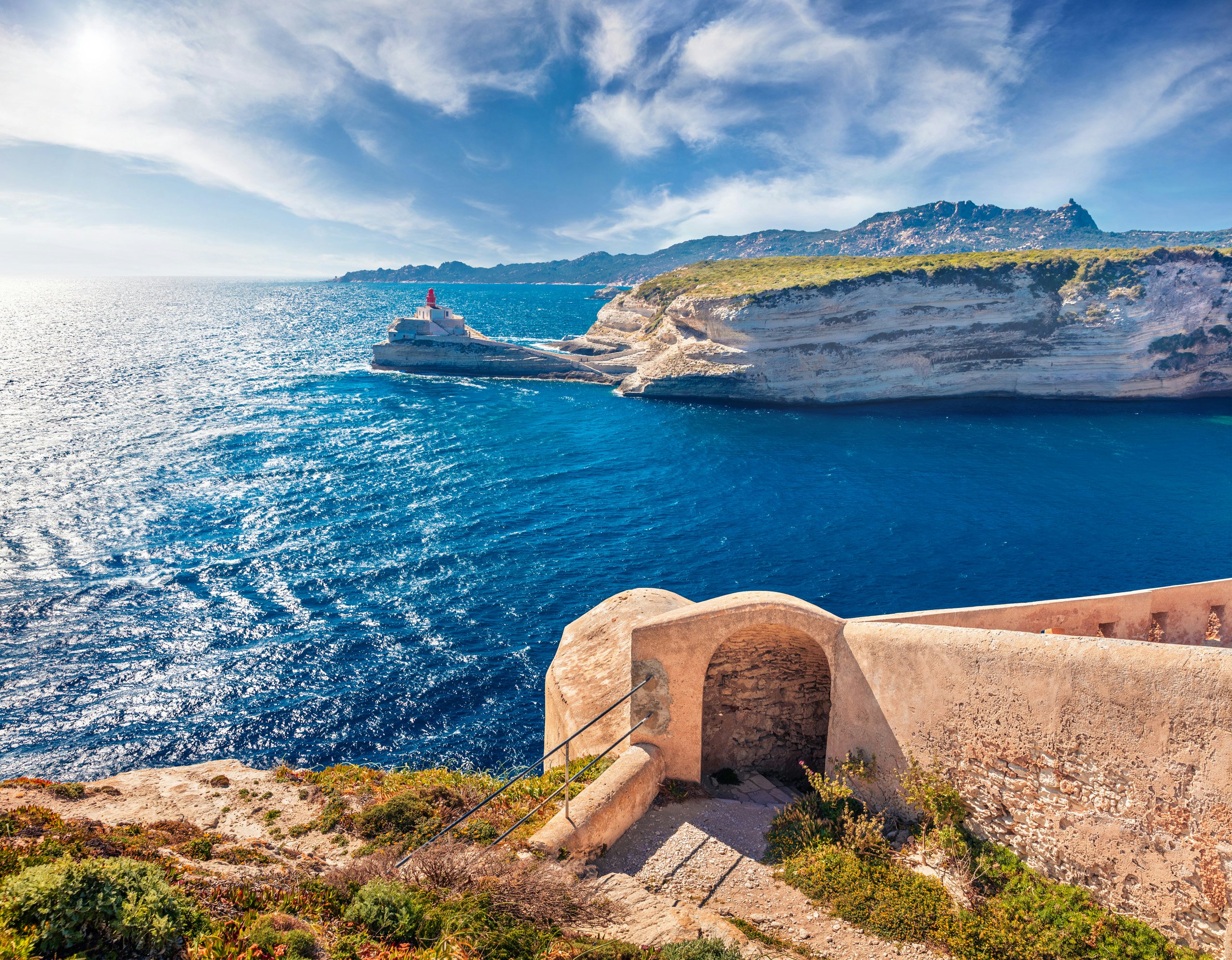 A cruise ship voyaging past sunlit cliffs viewed from a historic coastal pathway.