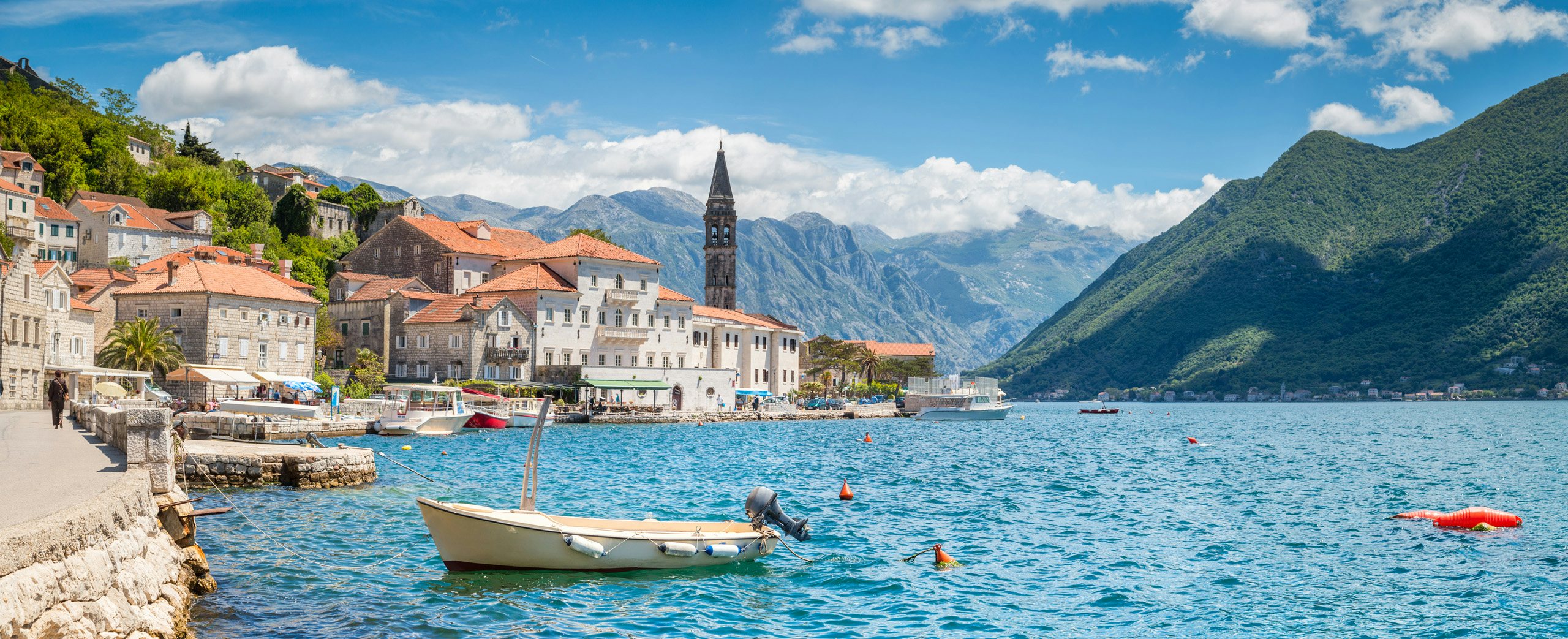 Serene Yacht Charter Destinations in Montenegro - N&J A small boat on the serene waters of Montenegro with a backdrop of mountains and historic buildings, ideal for yacht charter experiences by N&J.