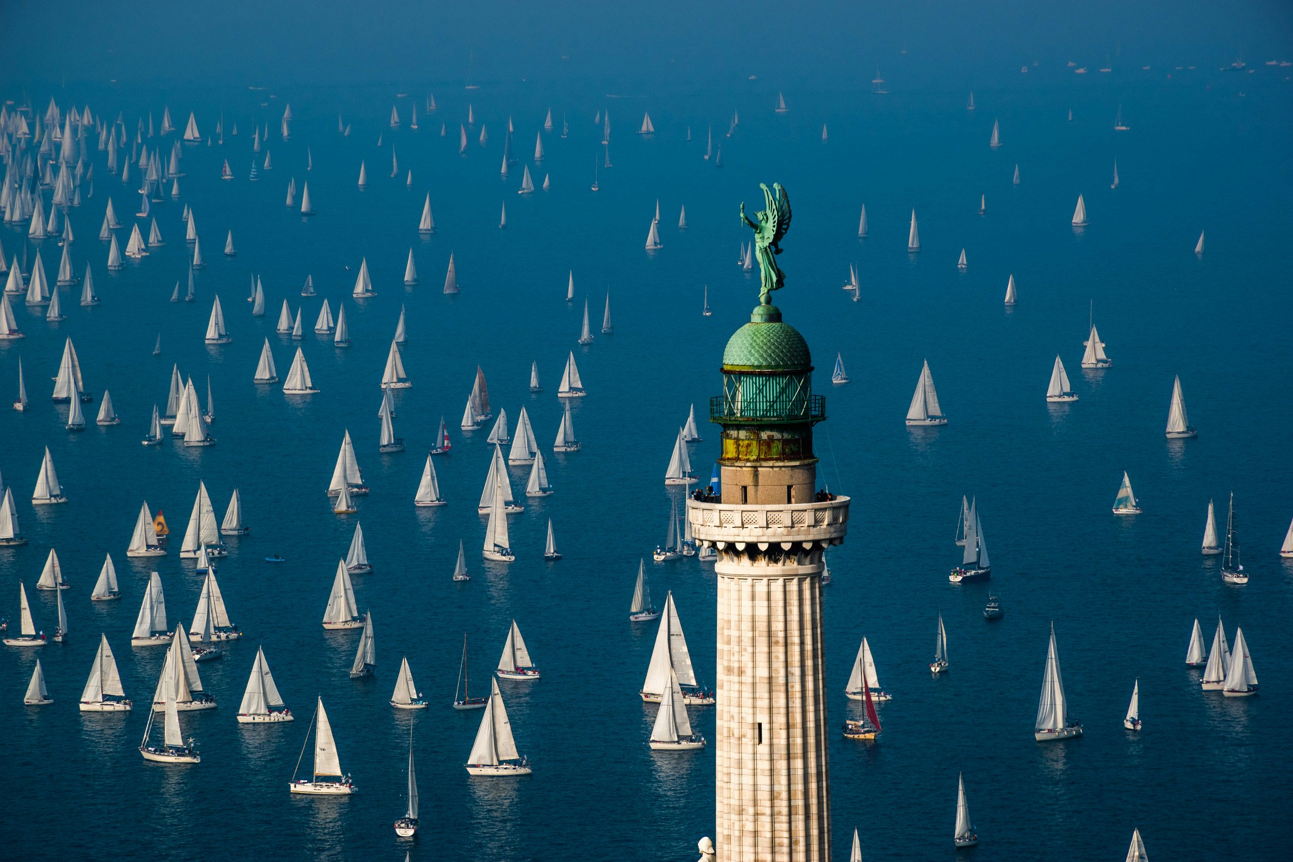 A lighthouse in Venice with a sea of sailboats in the background under a clear blue sky.