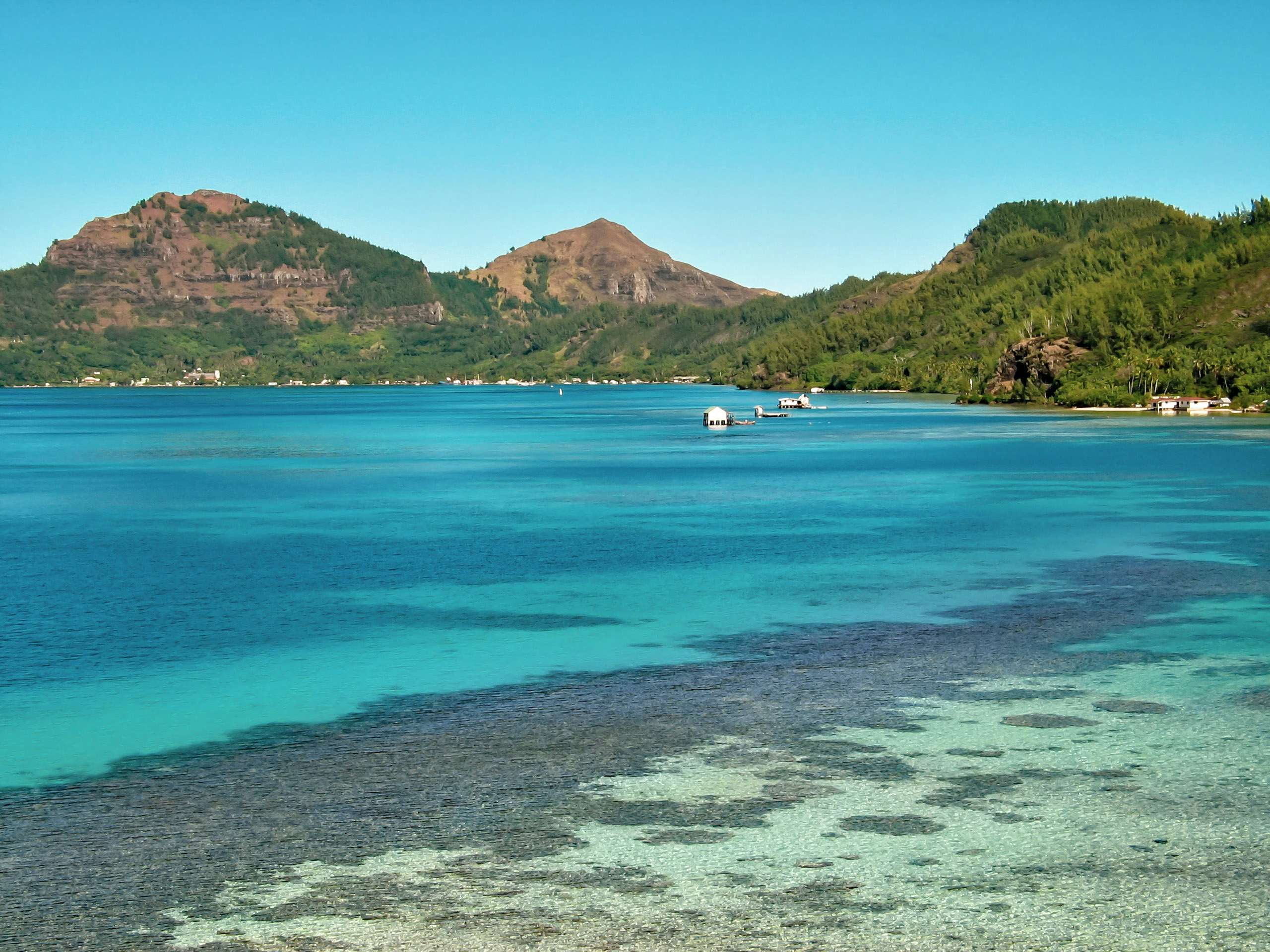 Tranquil coastal view of the Gambier Islands with clear blue waters and coral reefs in the foreground, anchored boats in the middle, and verdant hills in the background under a clear sky.