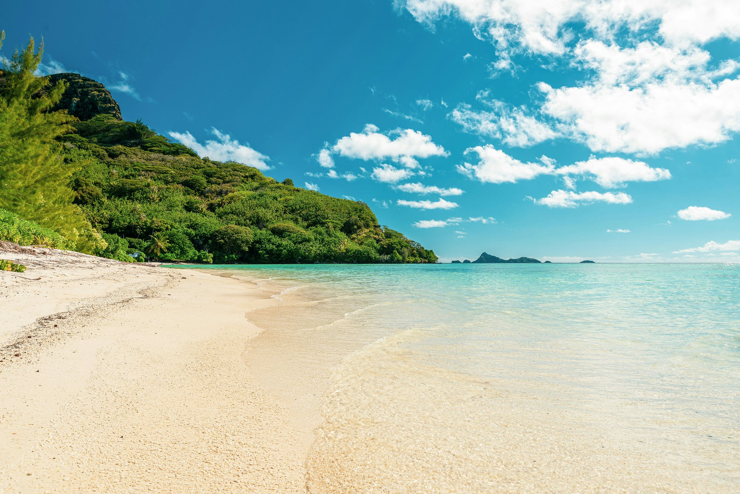 Pristine white sandy beach leading into transparent shallow waters with a verdant hillside on the Gambier Islands, and distant islets under a blue sky.