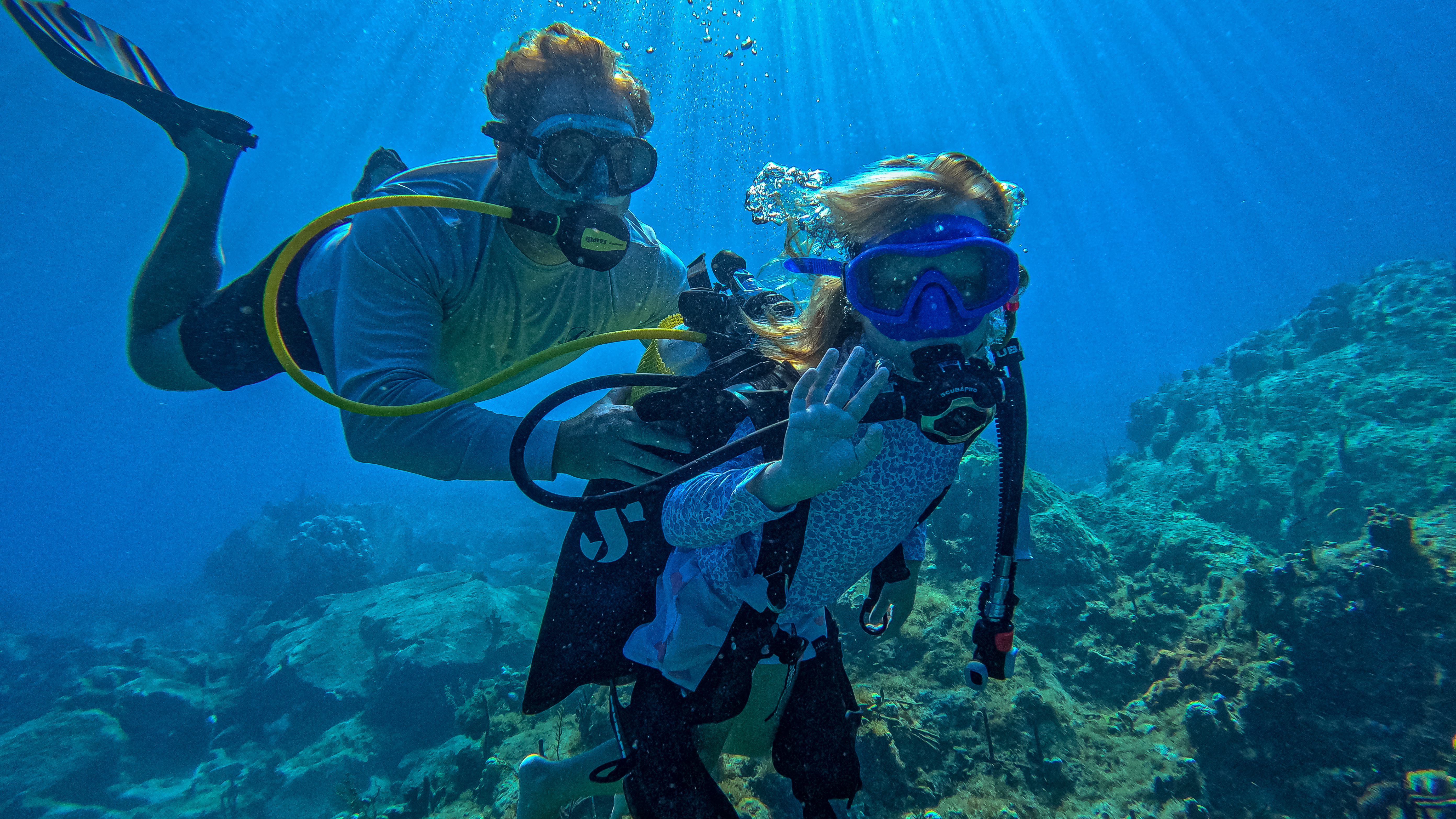 The image shows two people scuba diving in clear blue water. The person in the foreground appears to be assisting a child, both equipped with scuba gear including masks, regulators, and buoyancy compensators. Sunlight filters through the surface, illuminating the divers and the coral reef below. The divers are surrounded by the underwater environment, with coral and marine life visible in the background.