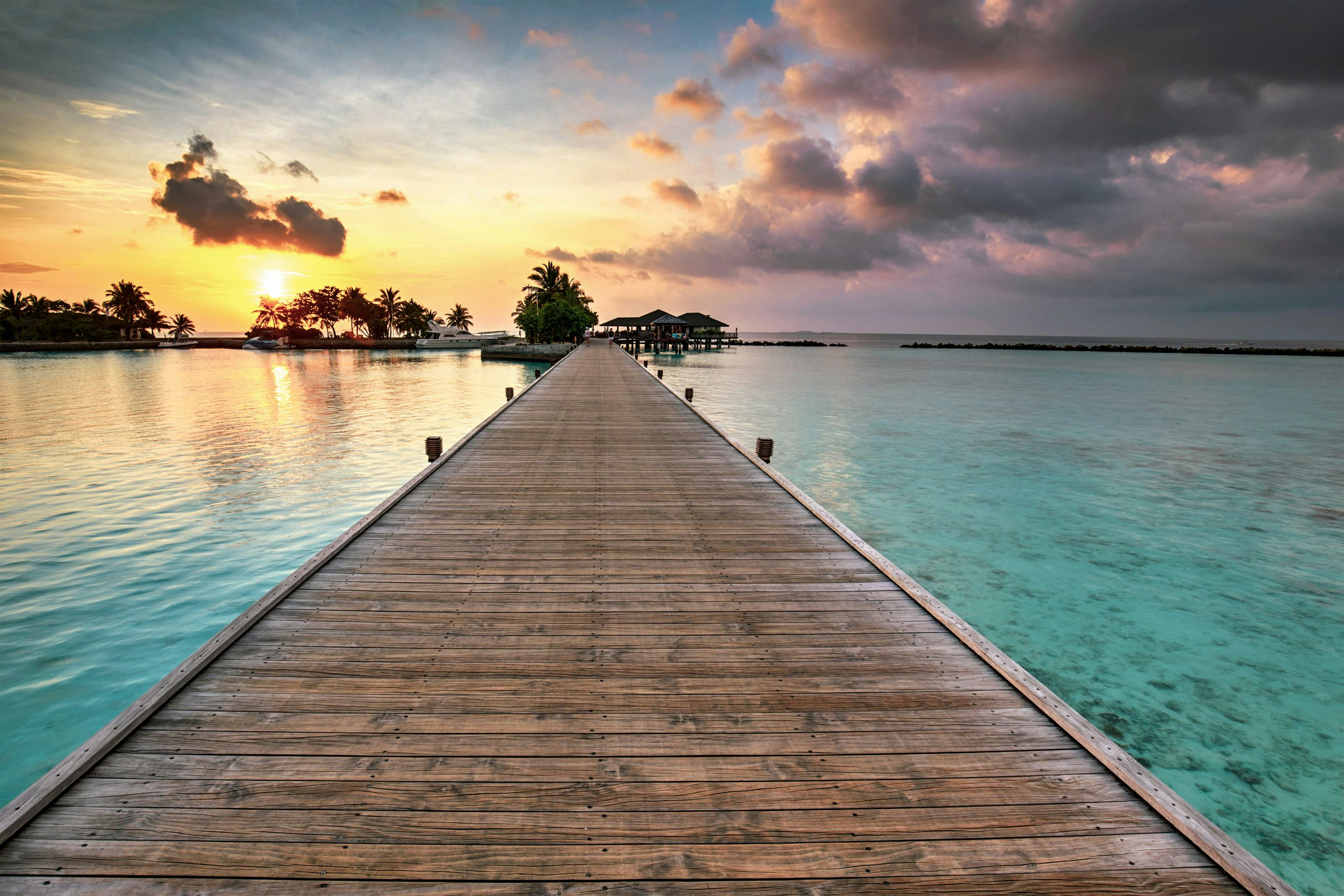 Gateway to Serenity - The Maldives Awaits Sunset colors the sky above a tranquil wooden jetty leading to an overwater resort in the Maldives, symbolizing an escape to paradise.