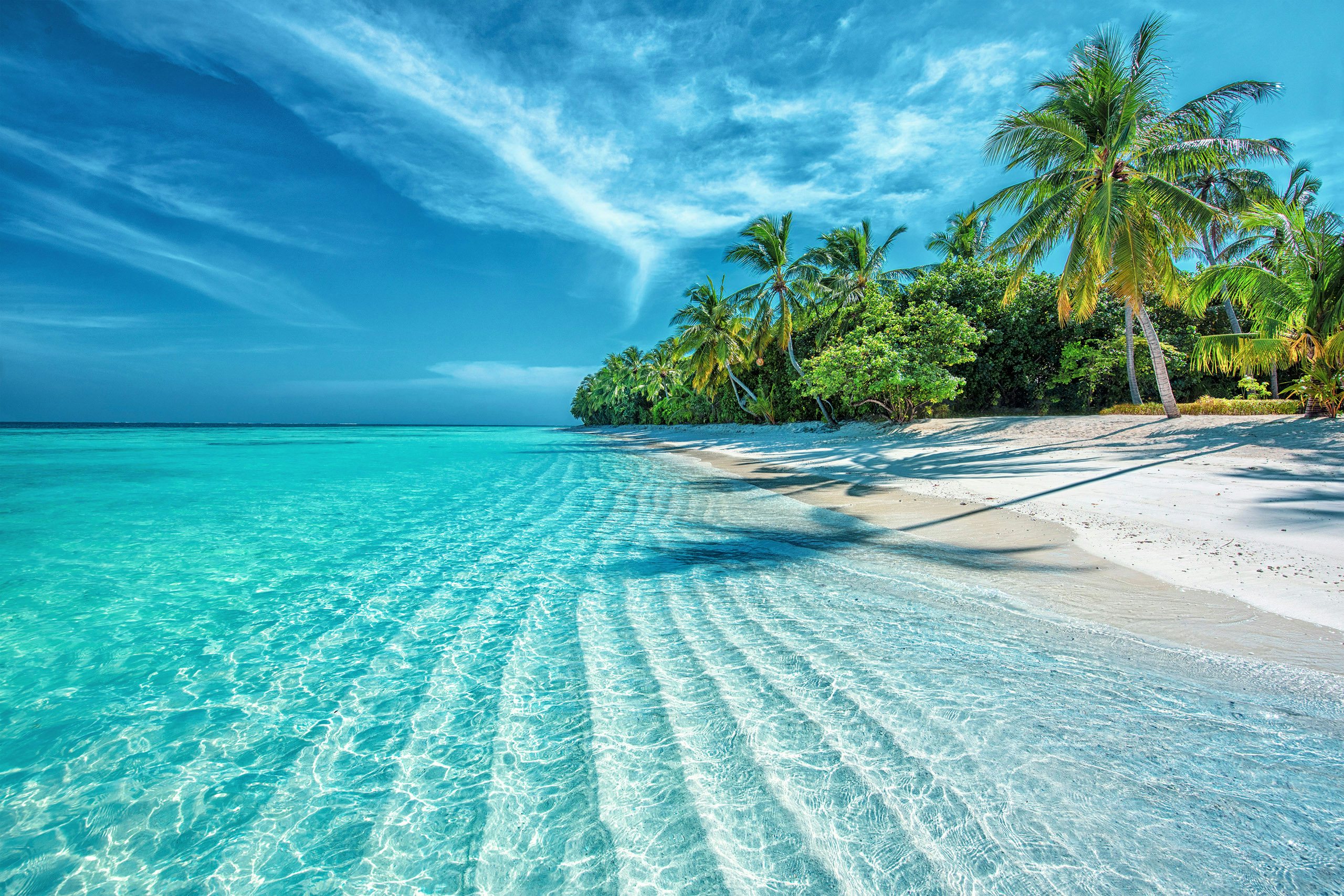 Crystal-clear shallow waters ripple over white sand, leading to a palm-fringed beach under a bright blue sky in the Maldives.