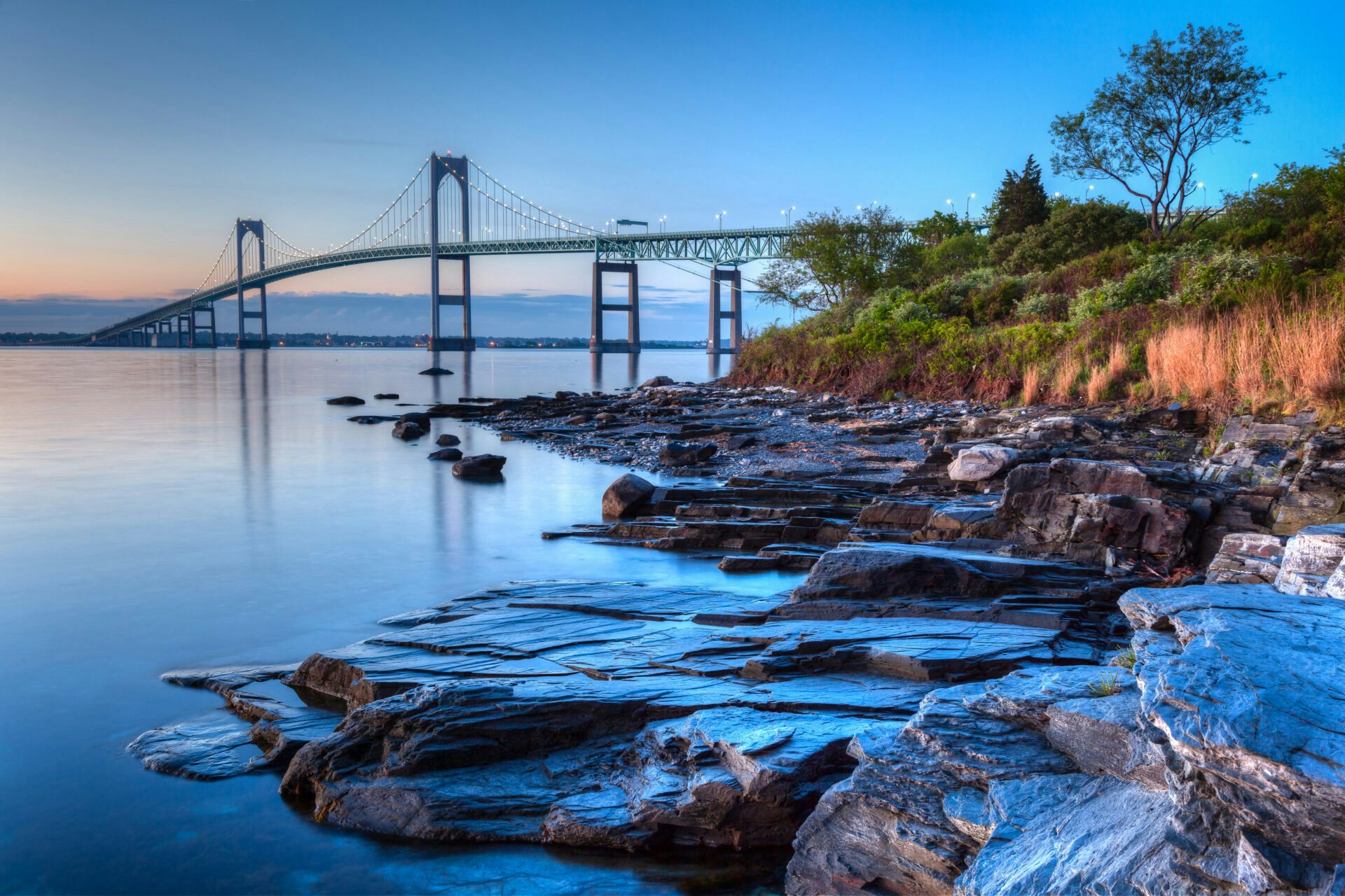 Newport Bridge Aglow: Sunset Serenity at the Newport Brokerage Show A captivating view of the Newport Bridge, with a rocky beach and lush green landscape, bathed in the warm hues of a Newport sunset during the Newport Brokerage Show.