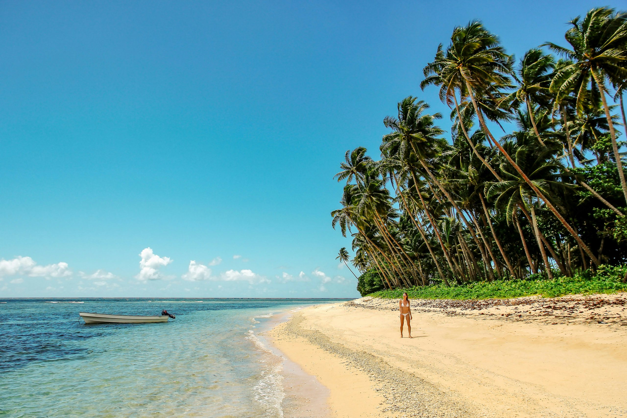 A solitary figure strolling on a palm-fringed beach in Fiji, with a canoe floating nearby.