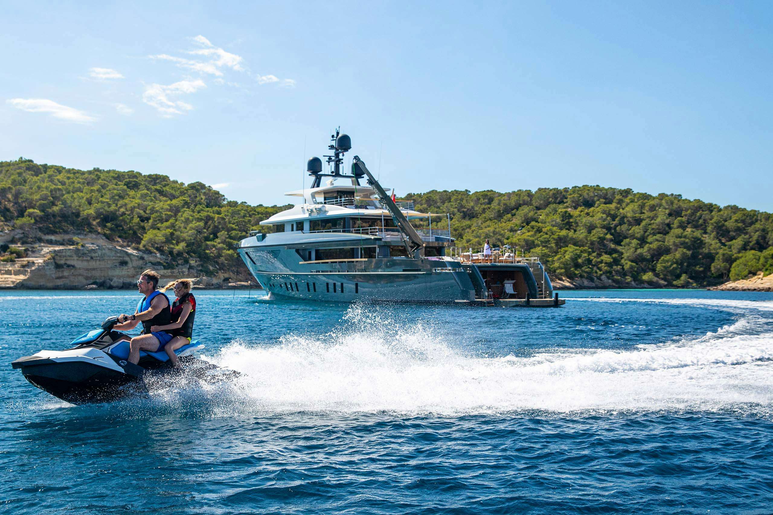 A couple enjoys jet skiing in front of a luxurious yacht anchored near a lush island, showcasing the exciting activities available during a yacht charter.