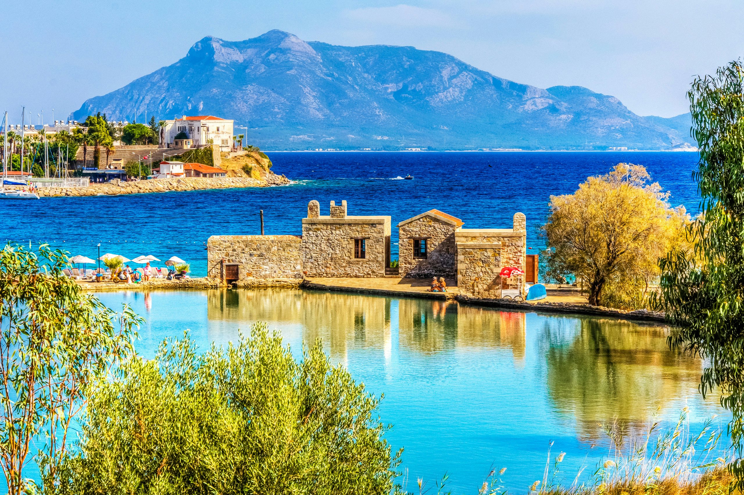 Tranquil Shores and Timeless Beauty - Discover Turkey by Yacht Quaint stone buildings by a serene lake with a mountainous backdrop and clear blue skies.