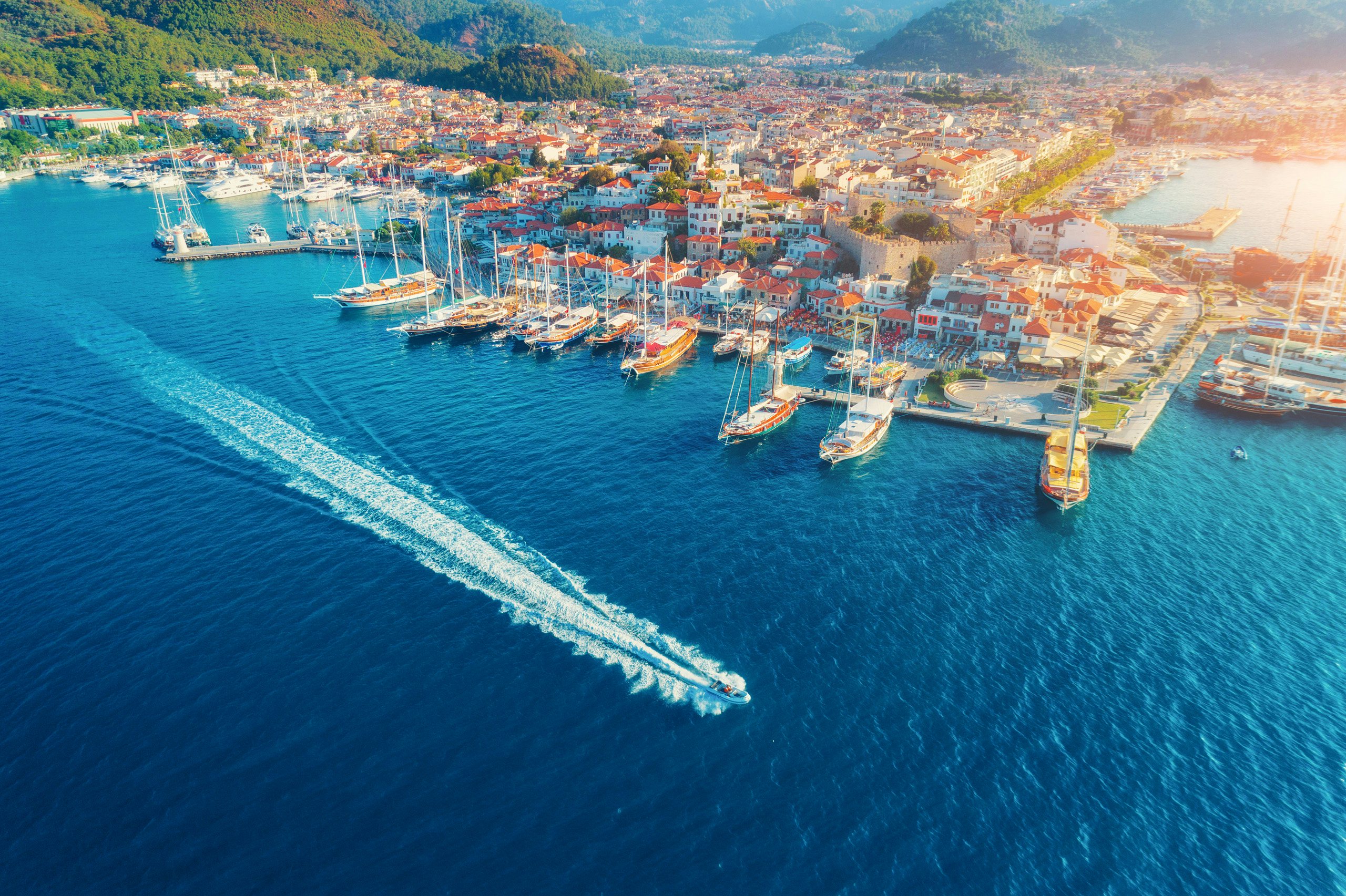 Aerial view of a coastal town with moored yachts and a prominent yacht leaving the harbor.