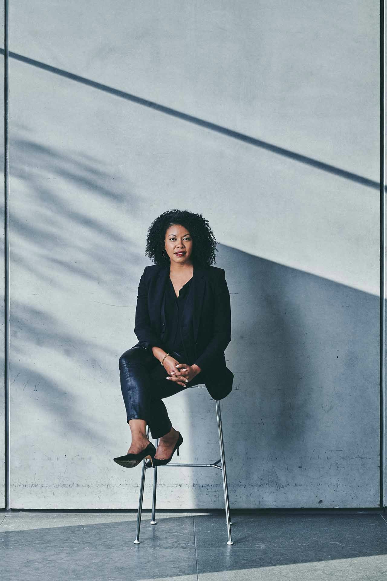 A woman in a black suit sitting on a chair in front of a grey concrete wall