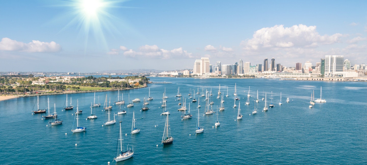 A picturesque view of numerous sailboats anchored outside the San Diego International Boat Show, with the San Diego coastline and skyline in the background, set against the backdrop of a sunny day and the sparkling blue sea.