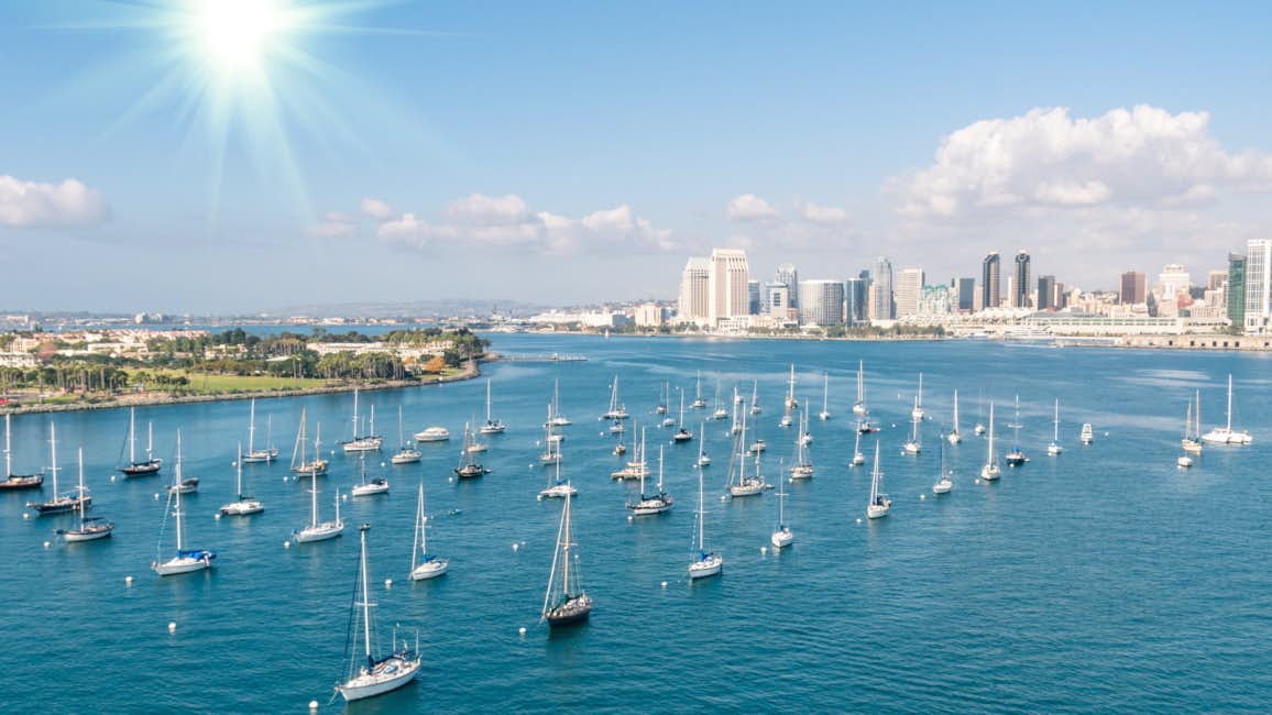 A picturesque view of numerous sailboats anchored outside the San Diego International Boat Show, with the San Diego coastline and skyline in the background, set against the backdrop of a sunny day and the sparkling blue sea.