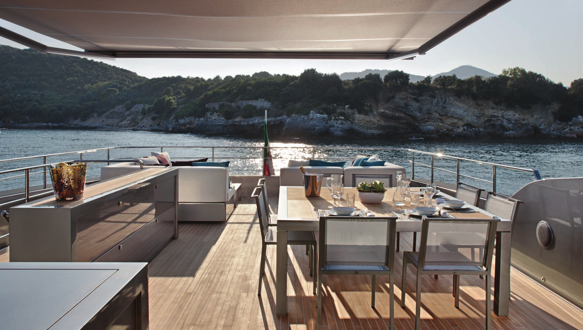 An elegant outdoor dining setup on the Sanlorenzo 40 Alloy yacht, overlooking calm waters and a lush coastline at sunset.