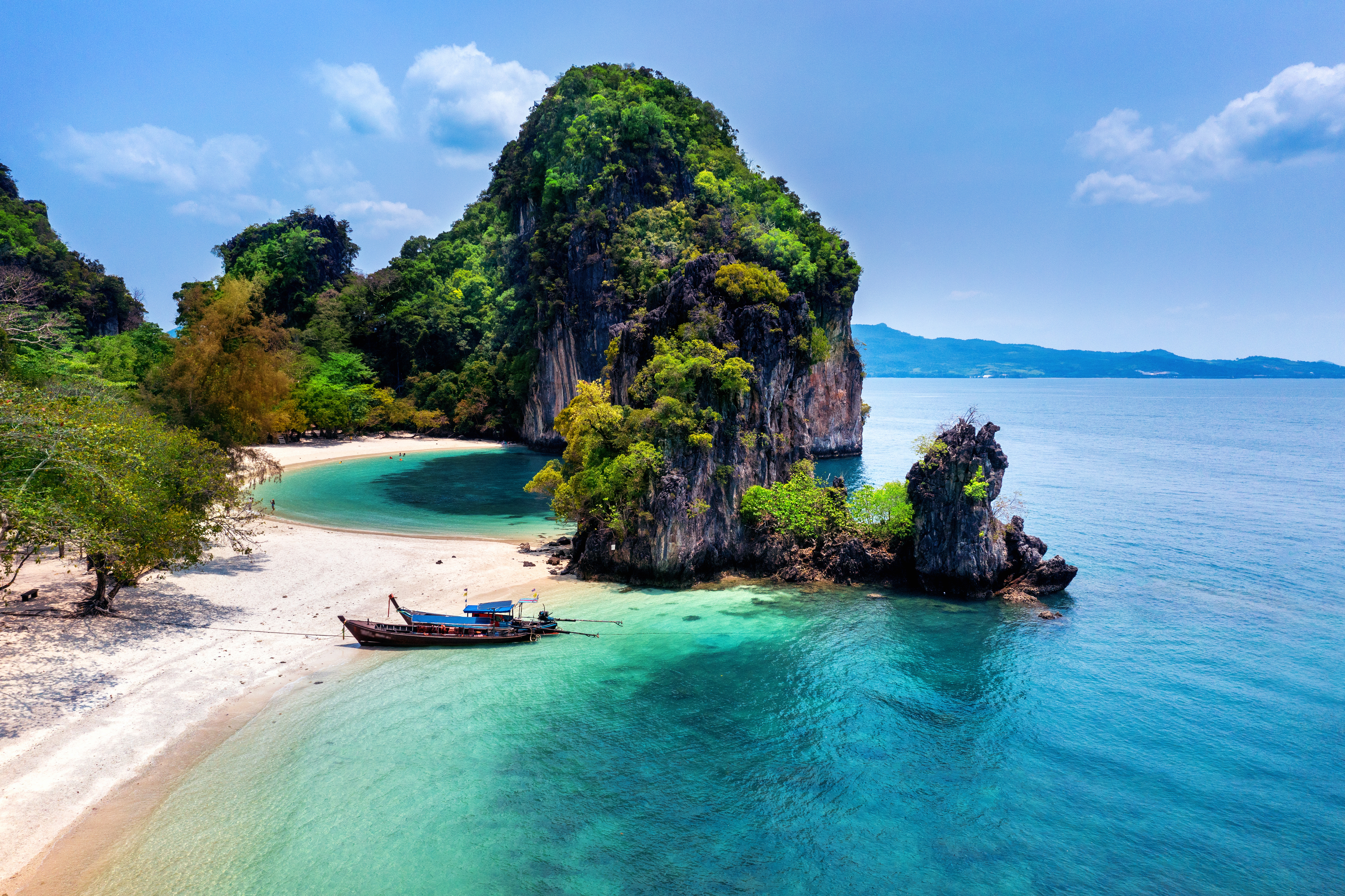 The image shows a beautiful tropical beach with crystal-clear turquoise water. A lush green island rises dramatically from the sea, covered in dense vegetation. A small boat rests on the sandy shore, and the sky is a clear blue with scattered clouds. The scene evokes a sense of tranquility and paradise.